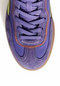 Zapatilla morada con material de ante y sintético, que presenta costuras naranjas, cordones texturizados y una suela de goma marrón. Detalle verde en el lateral.
