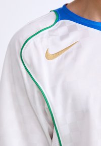 La camiseta deportiva blanca de Nike presenta un patrón gráfico texturizado, cuello azul, ribete verde y un logotipo bordado en dorado en la parte superior del pecho.