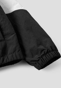 Sort-hvid jakkeærme med elastisk manchet og synlige syninger på lys grå baggrund.