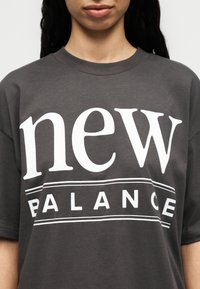 Γυναίκα που φοράει σκουρόγριμη μπλούζα με μεγάλο λευκό κείμενο "new BALANCE" στο κέντρο του στήθους.