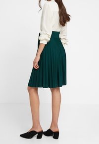 Grön veckad kjol med hög midja, tillverkad av mjukt tyg. Bärs med en ljusblommig blus och svarta sandaler med öppen tå.