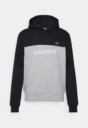 Musta ja harmaa huppari etutaskulla, "LACOSTE" teksti ja pieni vihreä krokotiililogo rinnassa.
