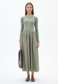 Lång, grön klänning med ribbat liv och ett samlat, flödande kjol. Modellen håller en brun väska; bär svarta platta sandaler. Minimalistisk design.