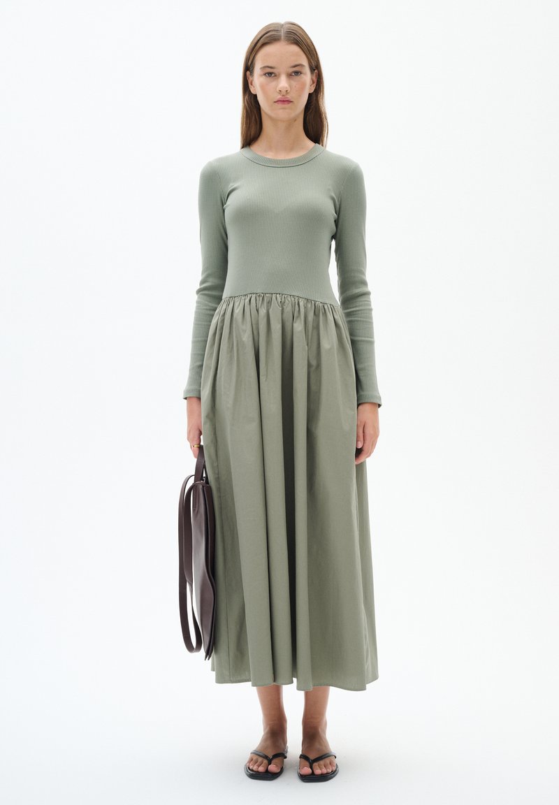 Lång, grön klänning med ribbat liv och ett samlat, flödande kjol. Modellen håller en brun väska; bär svarta platta sandaler. Minimalistisk design.