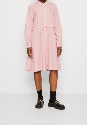 Kvinde iført en lyserød og hvidt lodretstribet knap-op kjole med lange ærmer, et taljebånd, sorte sokker og sorte loafers med guldkæder.