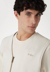 Helle beige Zip-Up-Weste mit einem gespaltenen Design. Verfügt über ein dezentes „GUESS“-Logo in passender Stickerei, das über einem cremefarbenen T-Shirt geschichtet ist.