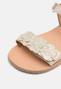 Silverfärgade lädersandaler med ett texturerat blommönster på remmen, en nude-färgad fotbädd och en slitstark grå yttersula. Närbilden framhäver detaljerna.