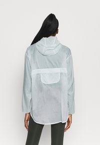 Chaqueta ligera y transpirable de color azul claro, con capucha, mangas largas y un bolsillo trasero. Presenta una textura suave y lisa y puños elásticos.