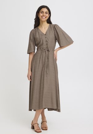 Femme souriante, portant une robe midi marron à petits pois blancs, manches mi-longues, devant boutonné, taille nouée, et sandales à talons marron.