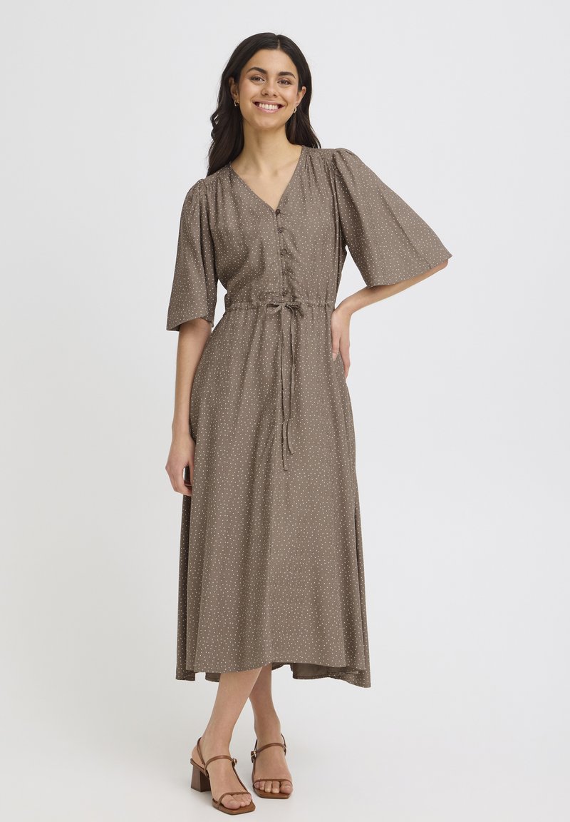 Femme souriante, portant une robe midi marron à petits pois blancs, manches mi-longues, devant boutonné, taille nouée, et sandales à talons marron.