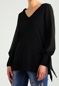 Schwarzer V-Ausschnitt-Pullover mit langen transparenten Ärmeln und gebundenen Manschetten, kombiniert mit bestickten blauen Jeans mit floralen Mustern.