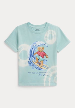 Svijetloplava majica s prstenovima tie-dye uzorka i grafikom medvjeda koji surfa na valovima, nosi crvenu majicu i zelene kratke hlače.