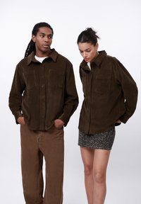 Twee personen dragen bruine corduroy shirts met borstzakken. Eén paar bruine broek en één rok met luipaardprint. Neutrale achtergrond.