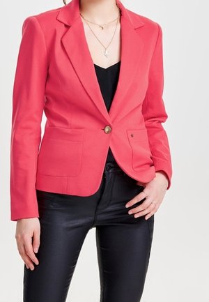 Femme portant un blazer rose corail sur un top noir et un pantalon en cuir noir, avec des colliers en or superposés et des bagues aux doigts.