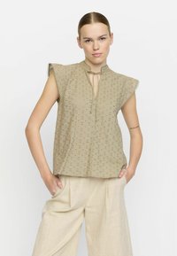 Beige blus med korta volangärmar, ögla-mönster, mandarin-krage och detalj med slips. Buren med ljusa, löst sittande byxor.