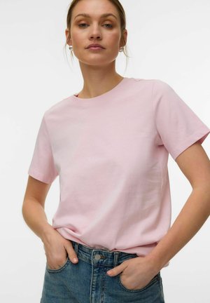 Woman wearing light pink short-sleeve t-shirt tucked into blue jeans, with hands in pockets against a plain white background.