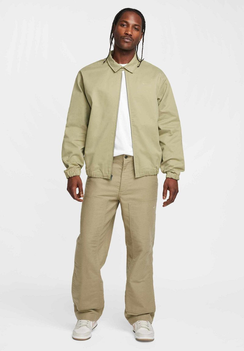 Chaqueta bomber verde claro sobre una camiseta blanca, combinada con pantalones beiges de corte holgado y zapatillas blancas. Diseño y ajuste simples y casuales.