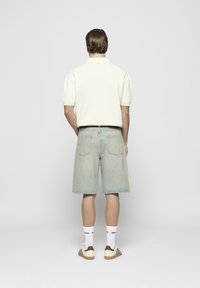 Polo de color beige claro, pantalones cortos de mezclilla en azul desvaído, calcetines blancos y zapatillas con acentos marrones. Vista desde atrás.