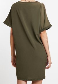 Robe vert olive à coupe décontractée, manches courtes et décolleté arrondi. Présente un tissu texturé et de subtils accents de camouflage.