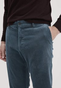 Blaue Cordhosen mit geripptem Muster, ausgestattet mit einem einzelnen Knopfverschluss und Seitentaschen, kombiniert mit einem dunklen Pullover.