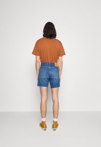 Brun bomullströja, blå denimshorts med fransad nederkant och gula sneakers. Bakifrån, visar detaljer på patchen vid midjan på shortsen.