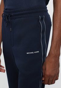 Tumši zilas sporta bikses, izgatavotas no mīksta auduma, ar aukliņu jostu un baltām sānu svītrām. Uz augšstilba redzams "MICHAEL KORS" logotips.