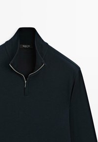 Zwarte zip-up pullover met hoge kraag. Gemaakt van zacht materiaal, met een ritssluiting aan de voorkant en subtiele schouderaccenten. Label zichtbaar bij de halslijn.