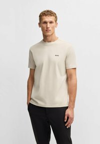 Beige kortærmet skjorte lavet af blødt stof, med rund hals og et lille sort "BOSS" logo på brystet. Sammen med sorte bukser.