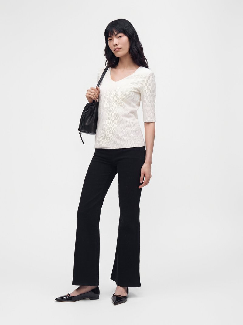 Femme aux longs cheveux noirs porte un haut blanc texturé, un pantalon flare noir, des ballerines noires et porte un sac à bandoulière noir.