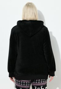 Sort fleece hoodie med hætte, blød tekstur, ribbede manchetter og kant; båret over mønstrede bukser med snefnug og rensdyr.