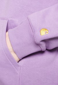 Handen instoppad i fickan på en ljuslila sweatshirt med ribbad mudd och en liten gul spiral-logotyp nära handleden.