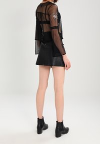Haut noir transparent à manches longues avec broderie florale, associé à une mini-jupe en cuir noir et des bottines, mettant en valeur une silhouette ajustée.
