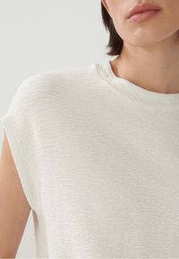 Bílý top s krátkým rukávem, vyrobený z texturované tkaniny s jemným vzorem a kulatým výstřihem. Jednoduchý design s minimálním množstvím švů.