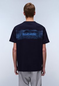 T-shirt noir à manches courtes avec un motif de montagne bleue et le texte "NAPAPIJRI geographic" au dos. Matériau en coton doux, coupe décontractée.