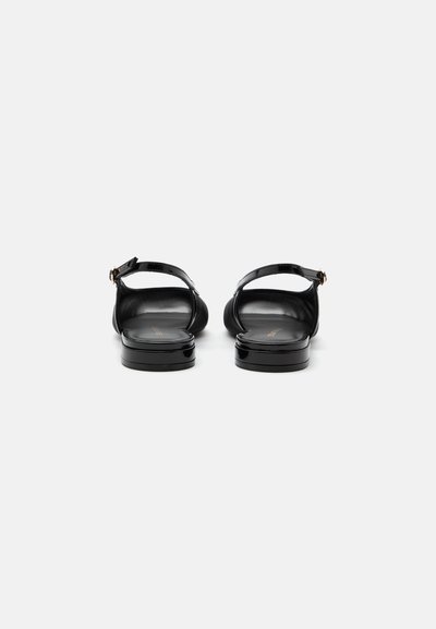 Paire de sandales plates en cuir verni noir avec des brides à la cheville et des boucles dorées, vues de dos sur fond blanc.