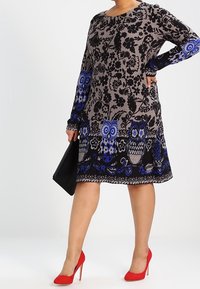 Robe à manches longues noire et grise avec motifs de hiboux et de fleurs en bleu et blanc. Portée avec des talons hauts rouges, tenant un sac à main noir.