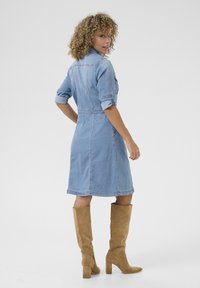 Denim kleita ar sarullētām piedurknēm, pieguļošu augšdaļu un A līnijas svārkiem; gaiši zila krāsa; apvienota ar brūnām zamša ceļgala zābakiem.