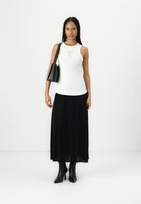 Weißes Tanktop mit Rundhalsausschnitt und Aufdruck, kombiniert mit einem fließenden schwarzen, plissierten Maxirock und schwarzen Stiefeletten mit Absatz. Schwarze Handtasche.