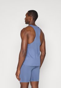 Modrý športový tielko s sieťovanou textúrou, dizajnom s prekríženými ramienkami a kontrastným stehom, v kombinácii s modrými šortkami, z pohľadu zozadu.