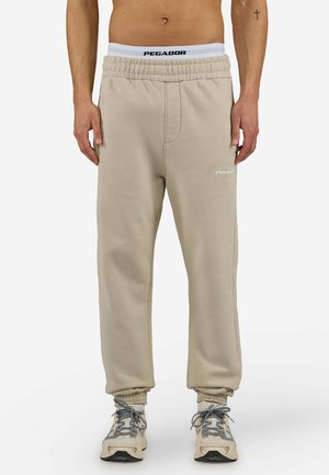 Man draagt beige joggingbroek met elastische tailleband en boorden, witte onderbroek zichtbaar, en beige en grijze sneakers.