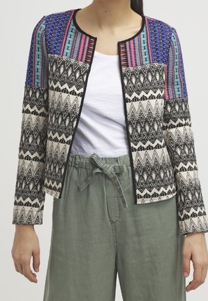 Vrouw draagt een meerkleurige jas met zwart-witte geometrische patronen over een wit overhemd en losse groene broek met een gestrikte taille.