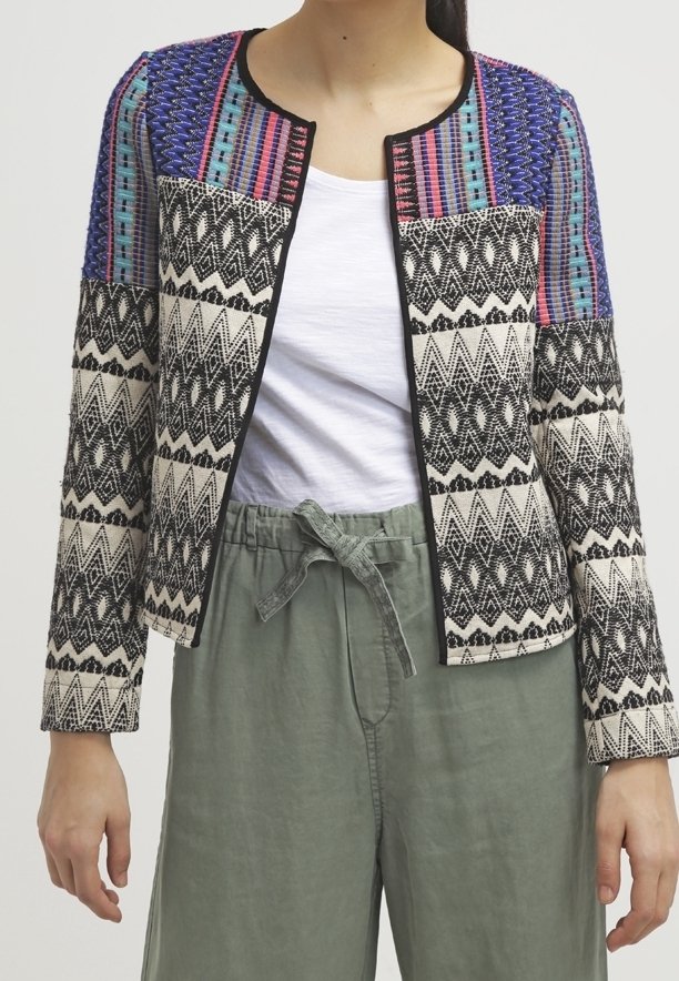 Femme portant une veste à motifs multicolores avec des motifs géométriques noirs et blancs sur une chemise blanche et un pantalon vert ample avec une taille nouée.