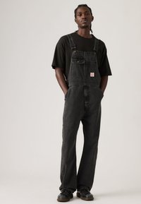 Νεαρός με κοτσίδες, φορώντας μαύρο μπλουζάκι, σκουρόχρωμο γκρι τζιν σαλόπετα και μαύρα παπούτσια, στέκεται με τα χέρια στις τσέπες μπροστά σε απλό φόντο.