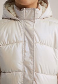Puffjacka i mjukt, ljust beige med hög krage. Har panelstygn och en fluffig innervadding för extra värme. Slät ytstruktur.