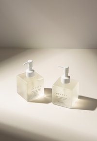 Due bottiglie pompa quadrate e traslucide di olio detergente. Le etichette dicono "NEEDLY Olio detergente profondo delicato". I pomelli bianchi e l'illuminazione soffusa mettono in risalto il design.