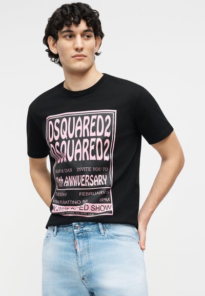 Jeune homme aux cheveux bouclés portant un t-shirt noir Dsquared2 avec un grand texte rose et un jean bleu clair, debout devant un fond uni.