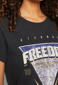 Camiseta de algodón negra con estampado gráfico que presenta la palabra "LIBERTAD" en un diseño triangular desgastado en blanco y morado, escote redondo y mangas cortas.