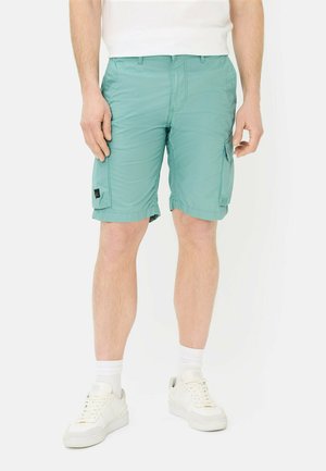 Man draagt lichtgroene cargoshorts, witte sokken, witte sneakers en een wit T-shirt, staand tegen een effen achtergrond.