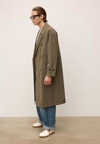Personne avec les cheveux lissés en arrière et des lunettes de soleil, portant un trench-coat vert olive surdimensionné, un jean bleu retroussé et des baskets blanches, se tenant de profil.