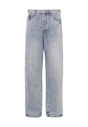 Femme portant un haut vert sans manches, un jean ample bleu clair, des chaussures noires à bout pointu, une casquette foncée et des lunettes de soleil, debout les bras croisés.